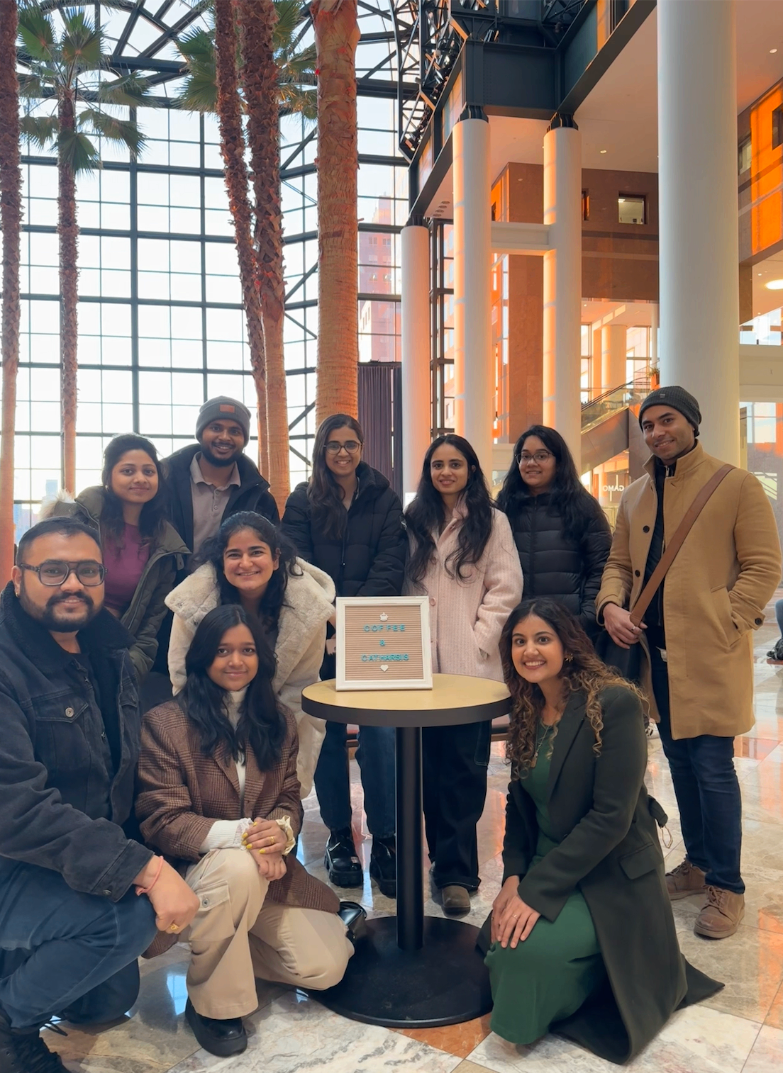 A group of our first Coffee & Catharsis attendees gathered around a small table with a letterboard sign that reads Coffee & Catharsis.