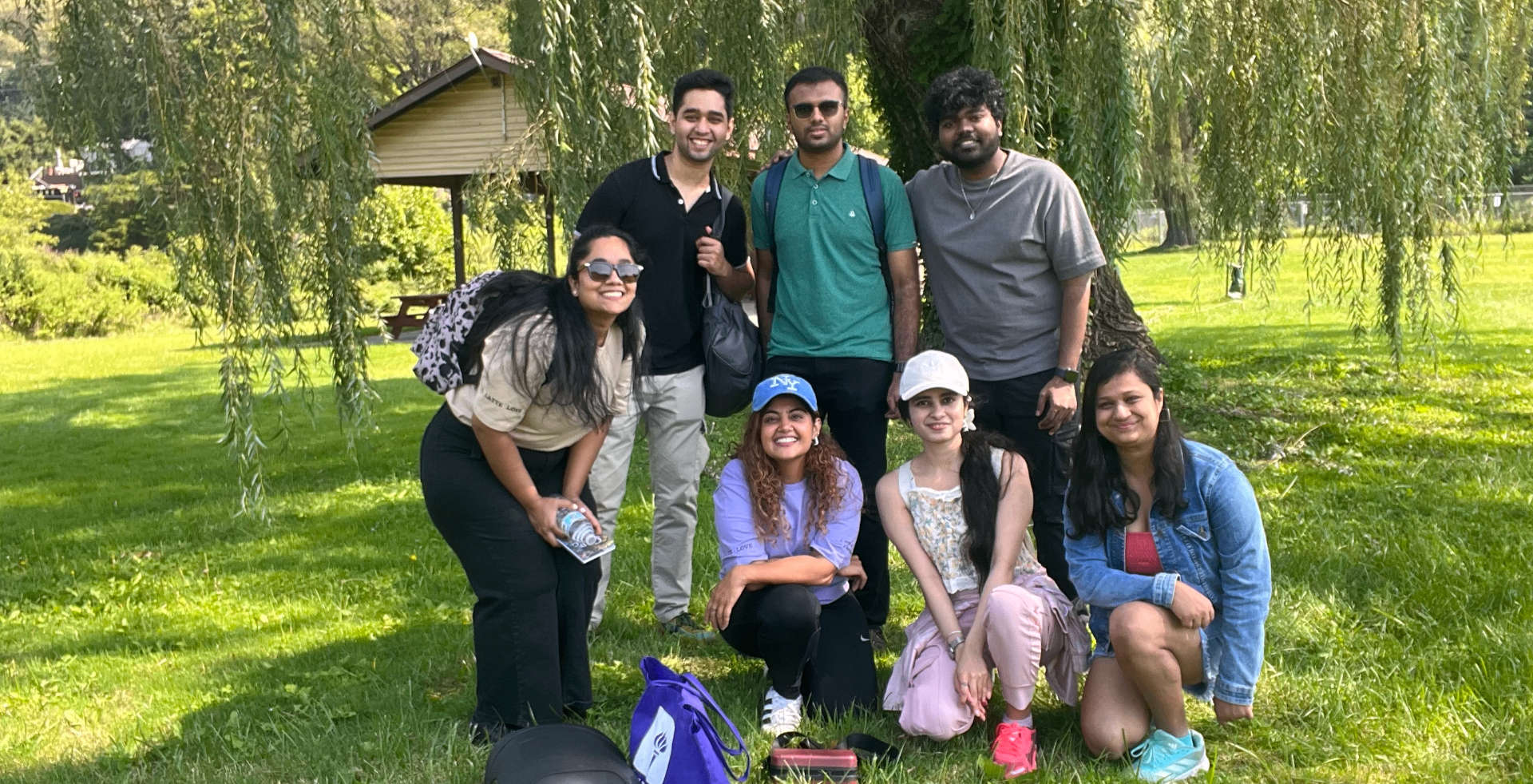 A group of friends smiling under a willow tree on a sunny day, outdoors together.
